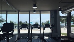 Une salle de sport équipée de tapis de course et de bancs d’exercices, de grandes fenêtres offrant une vue sur des palmiers et un parking.