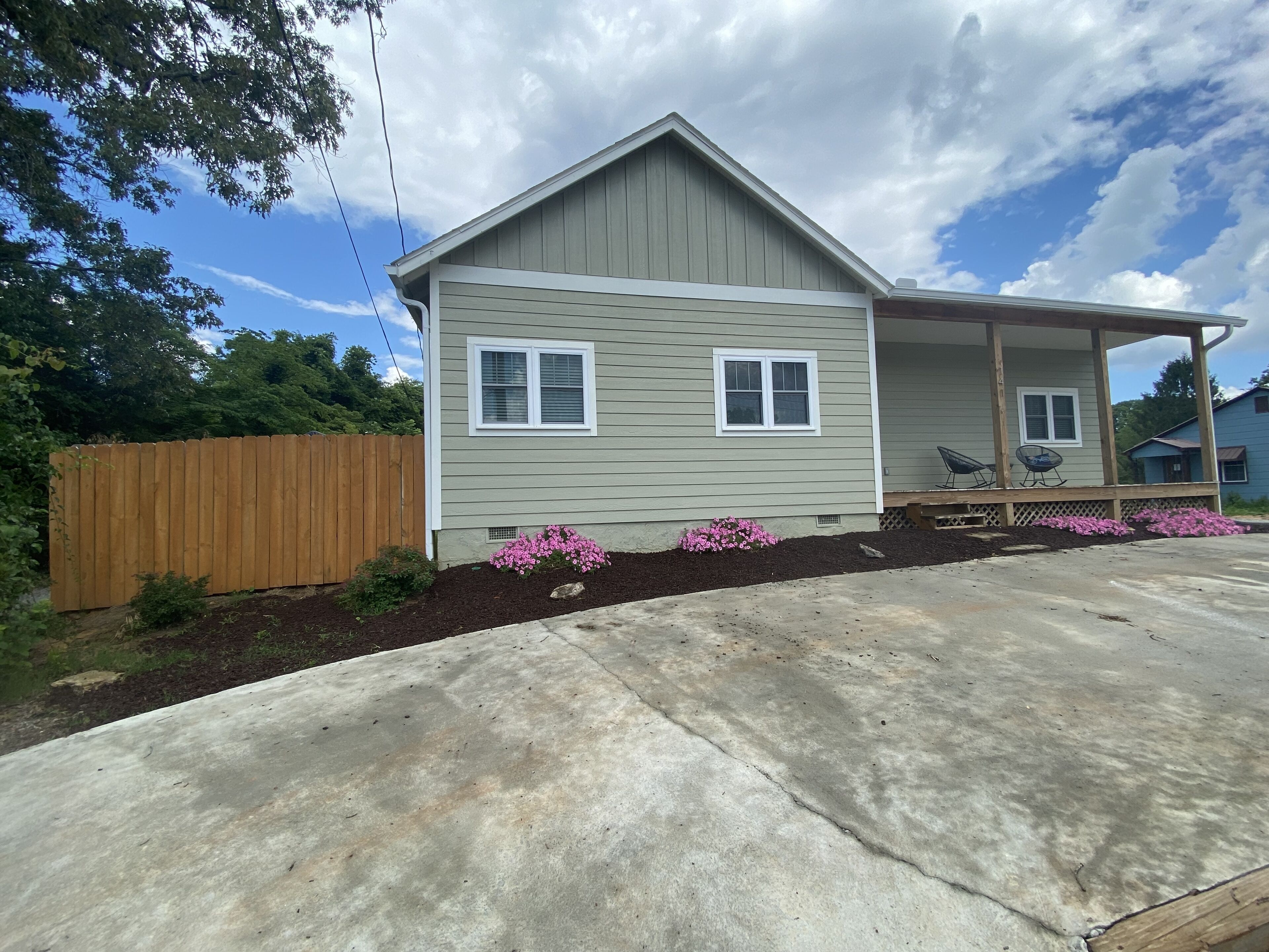 A single-story house with a porch, a wooden fence, and a garden with pink flowers.