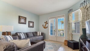 A living room with a grey sofa, a coffee table, and a TV stand. There is a door with a glass panel and a window with blinds. - Island Oasis! 1st floor! One block to the beach! (Corpus Christi)