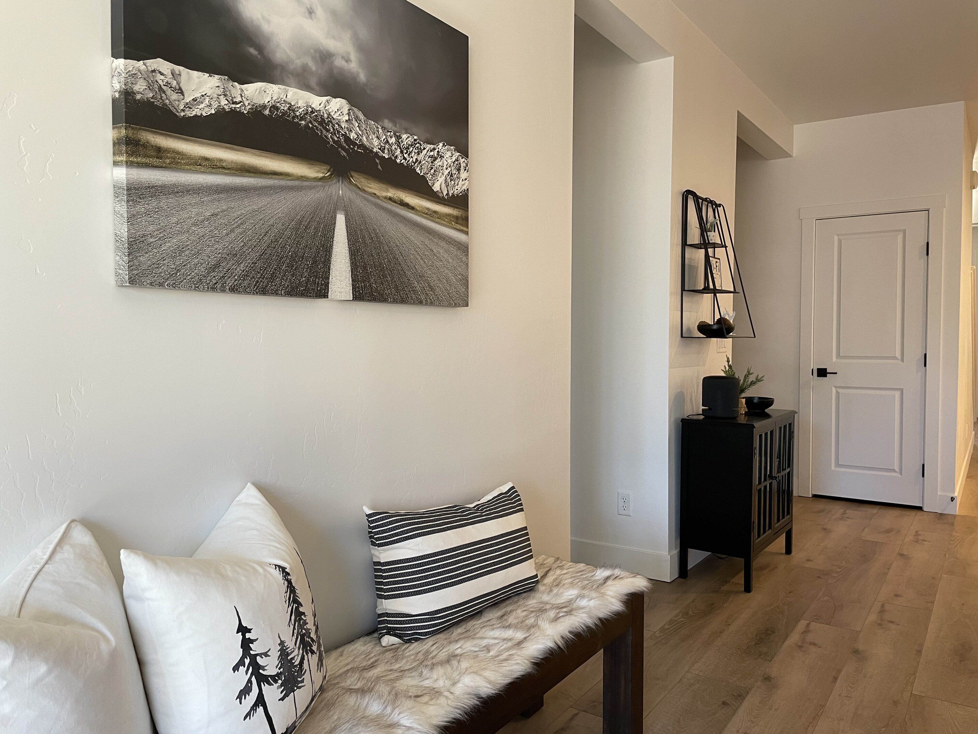 Een woonkamer met een bank, kussens, een houten bankje, een schilderij, een console tafel en een deur.