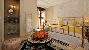Habitació d'hotel amb un llit, una taula rodona amb fruita, un otomà petit, un mirall i un bany amb un lavabo i un armari.