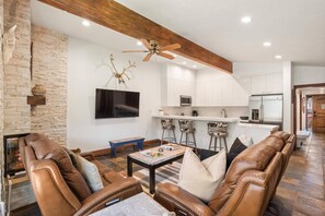 Una sala de estar moderna con sofás de cuero, chimenea, televisor de pantalla plana y una cocina con armarios blancos y taburetes de bar.