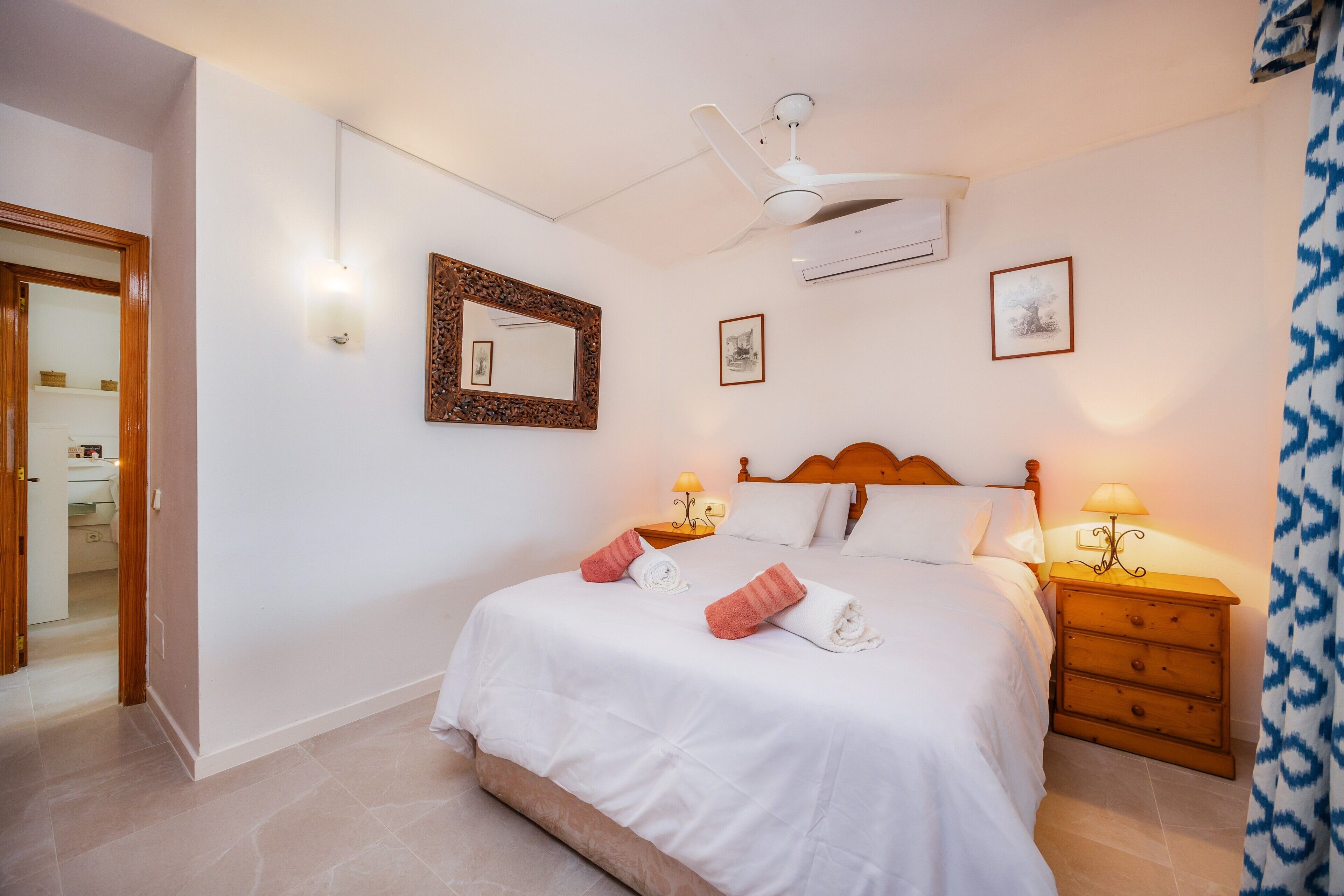 A bedroom with a bed, bedside tables, a ceiling fan, wall-mounted air conditioning, framed pictures, and a wooden door.
