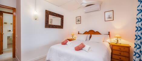 A bedroom with a bed, bedside tables, a ceiling fan, wall-mounted air conditioning, framed pictures, and a wooden door.