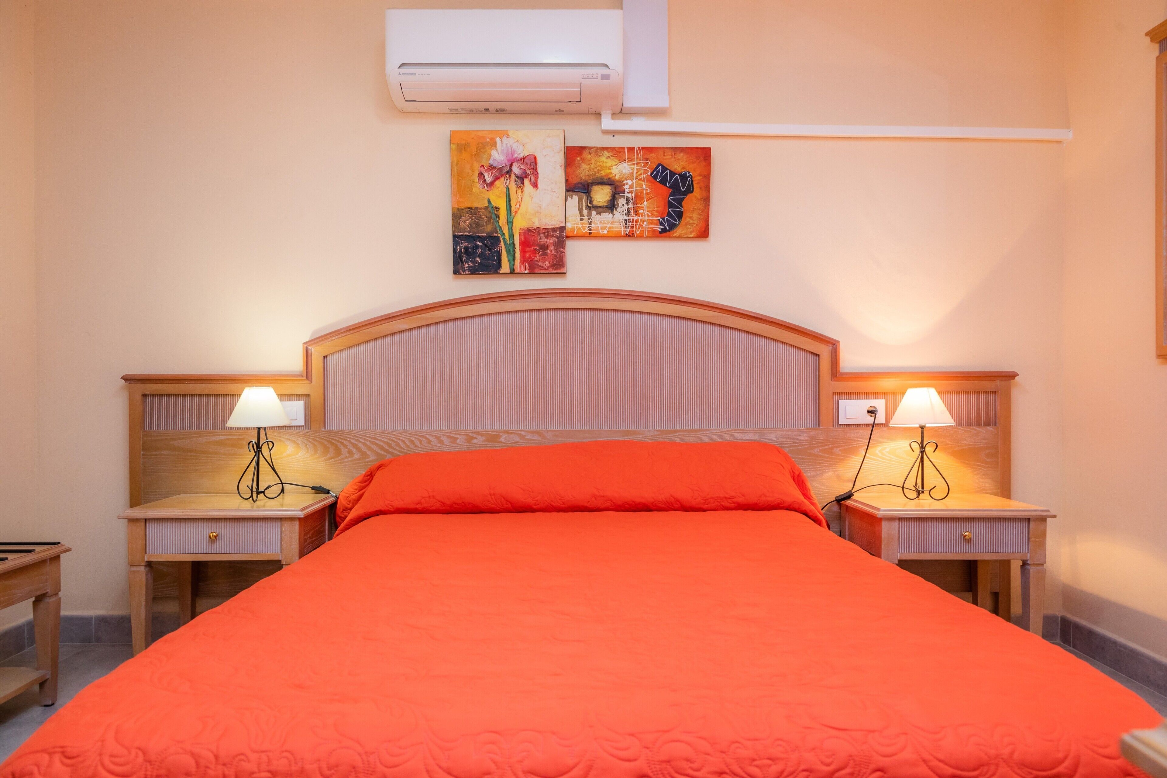 A hotel room with a bed, two bedside tables with lamps, an air conditioner, and two wall-mounted artworks.