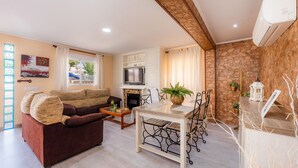 A living room with a sofa, coffee table, dining table, and chairs. There is a fireplace, kitchen area, and a stone wall. - "Can Caragol" Holiday Apartment with Private Terrace, A/C & WiFi (Cala Rajada)