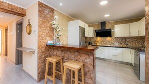 A modern kitchen with a stone accent wall, wooden stools, a built-in bar, and white cabinetry. - "Can Caragol" Holiday Apartment with Private Terrace, A/C & WiFi (Cala Rajada)