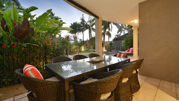 Un coin repas avec une table en verre et des chaises en osier, entouré de plantes tropicales et offrant une vue sur les palmiers.