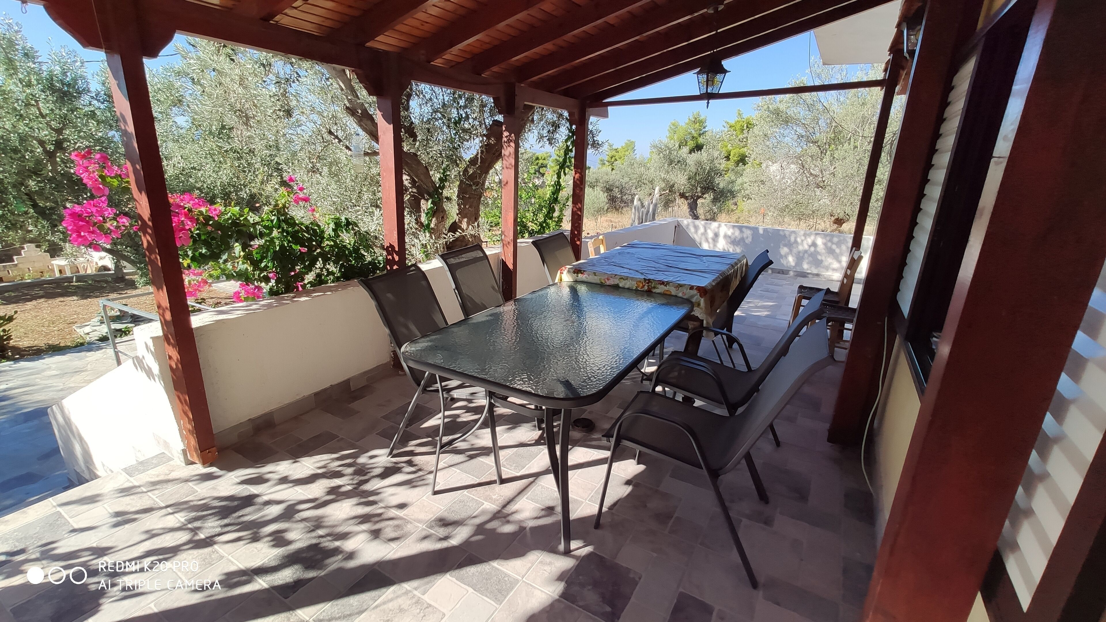 Un patio al aire libre con una mesa y sillas de vidrio, rodeado por una pérgola y vistas a árboles y flores.