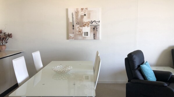 Een moderne woonkamer met een glazen salontafel, witte stoelen, een zwarte leren bank met een blauw kussen, en een abstract kunstwerk aan de muur.