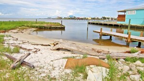 A beach with a wooden dock extending into calm waters, a sandy shore with driftwood, and a small blue building with a balcony.