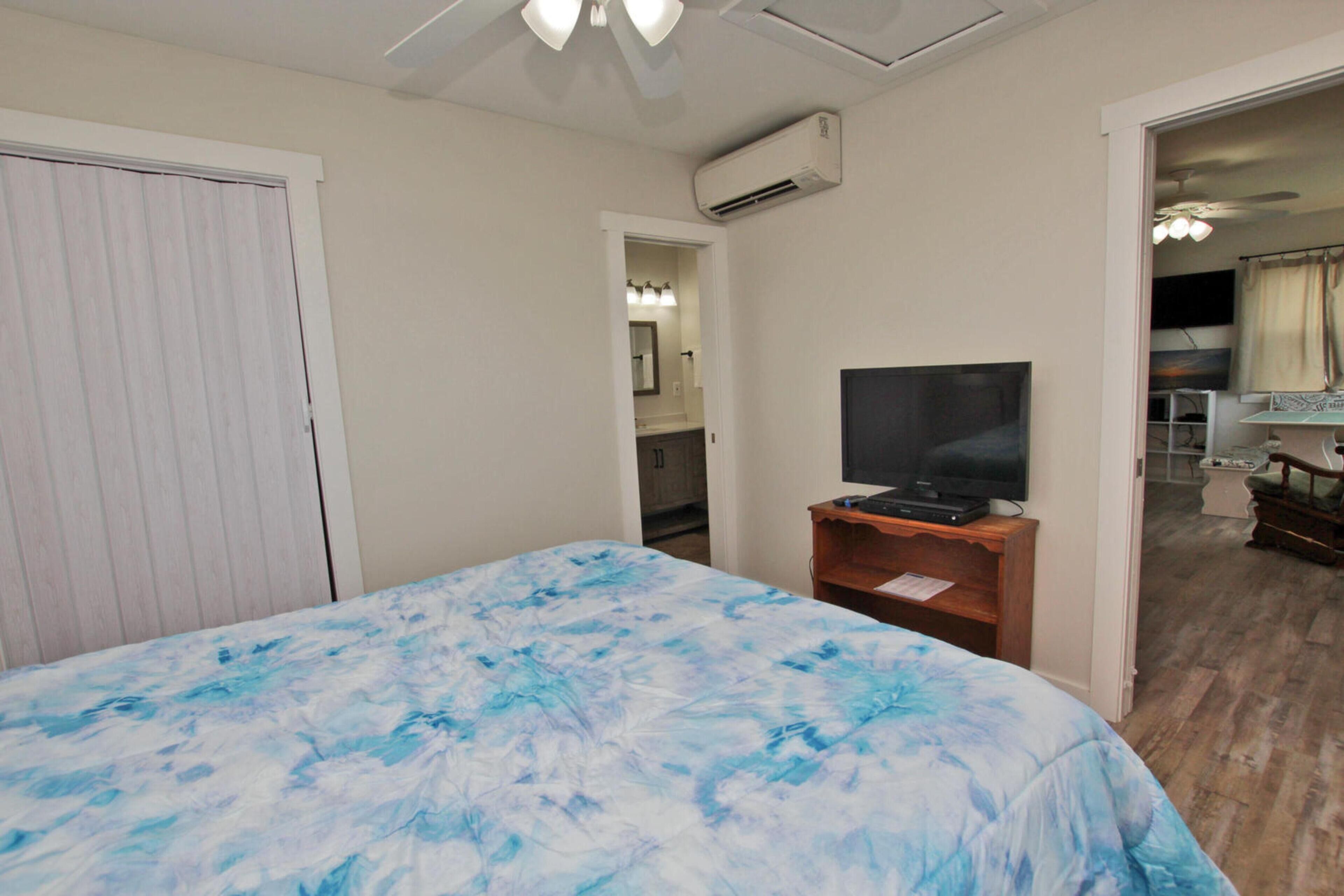 Un dormitorio con una cama, un televisor sobre un soporte de madera, un equipo de aire acondicionado y una puerta que da a otra habitación.