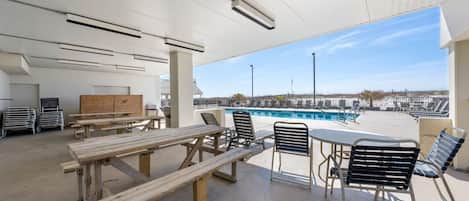 En utomhusplats med en uteplats, picknickbord och stolar, en pool i bakgrunden och fri utsikt över stranden.