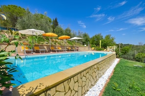Piscina com espreguiçadeiras e guarda-sóis, cercada por vegetação e um muro de pedra.