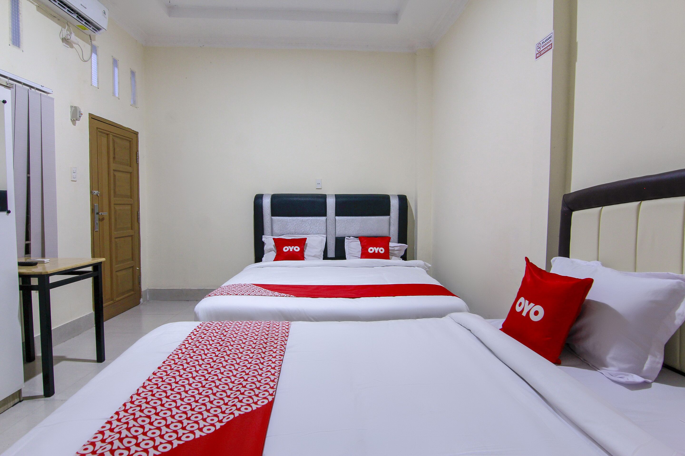 A hotel room with two beds, each with a red and white bedspread, a wooden door, a small table, and a wall-mounted air conditioner.