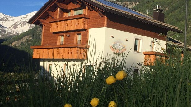 A wooden chalet with a balcony, surrounded by mountains and alpine vegetation.