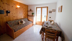 A cozy room with a wooden sofa, a dining table with chairs, a small kitchen area, and a sliding glass door leading to an outdoor area.