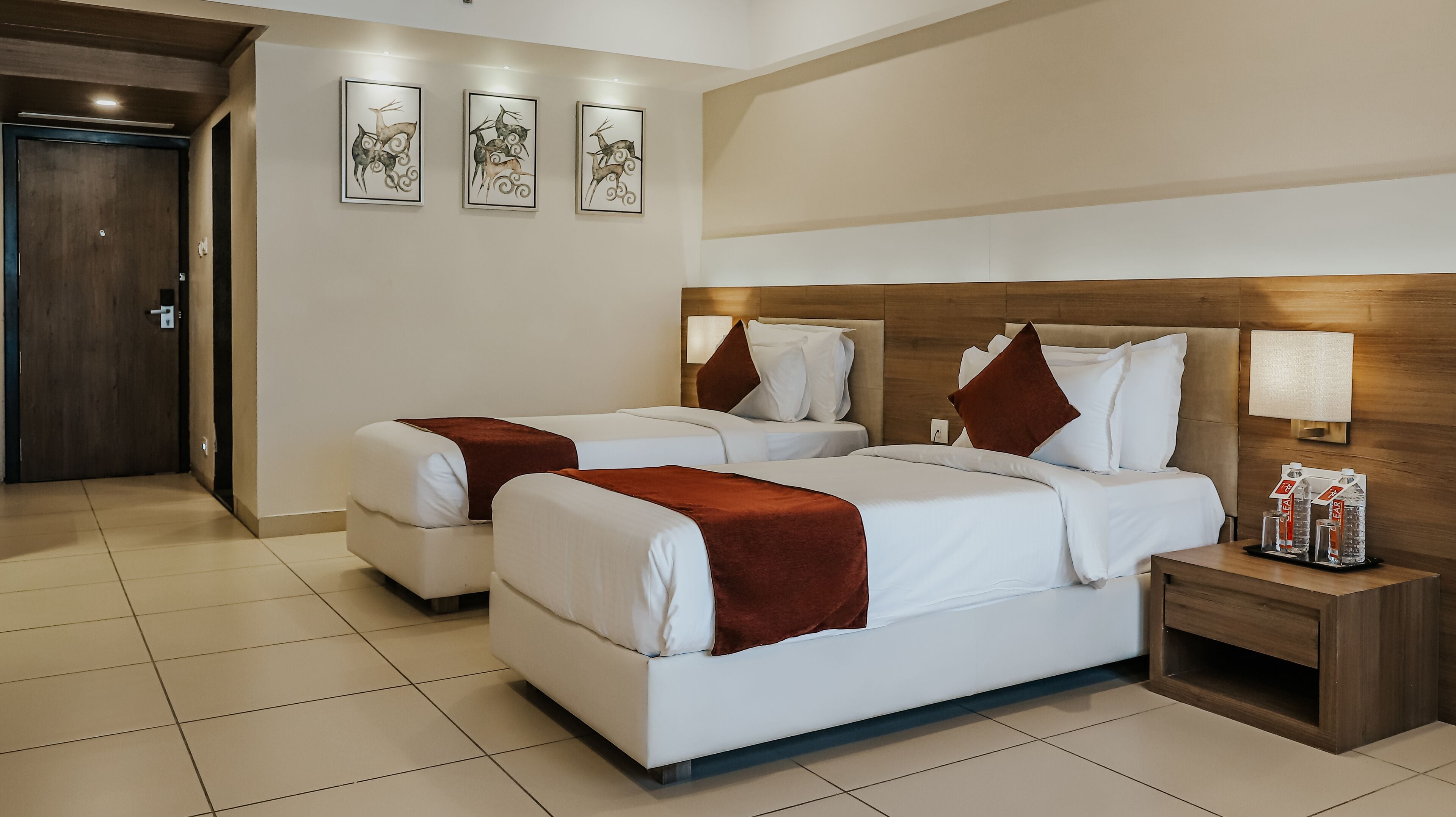 Une chambre d’hôtel avec deux lits, une tête de lit en bois, du linge de lit blanc, un plaid marron, une table de chevet avec des bouteilles d’eau et des œuvres d’art encadrées accrochées au mur.