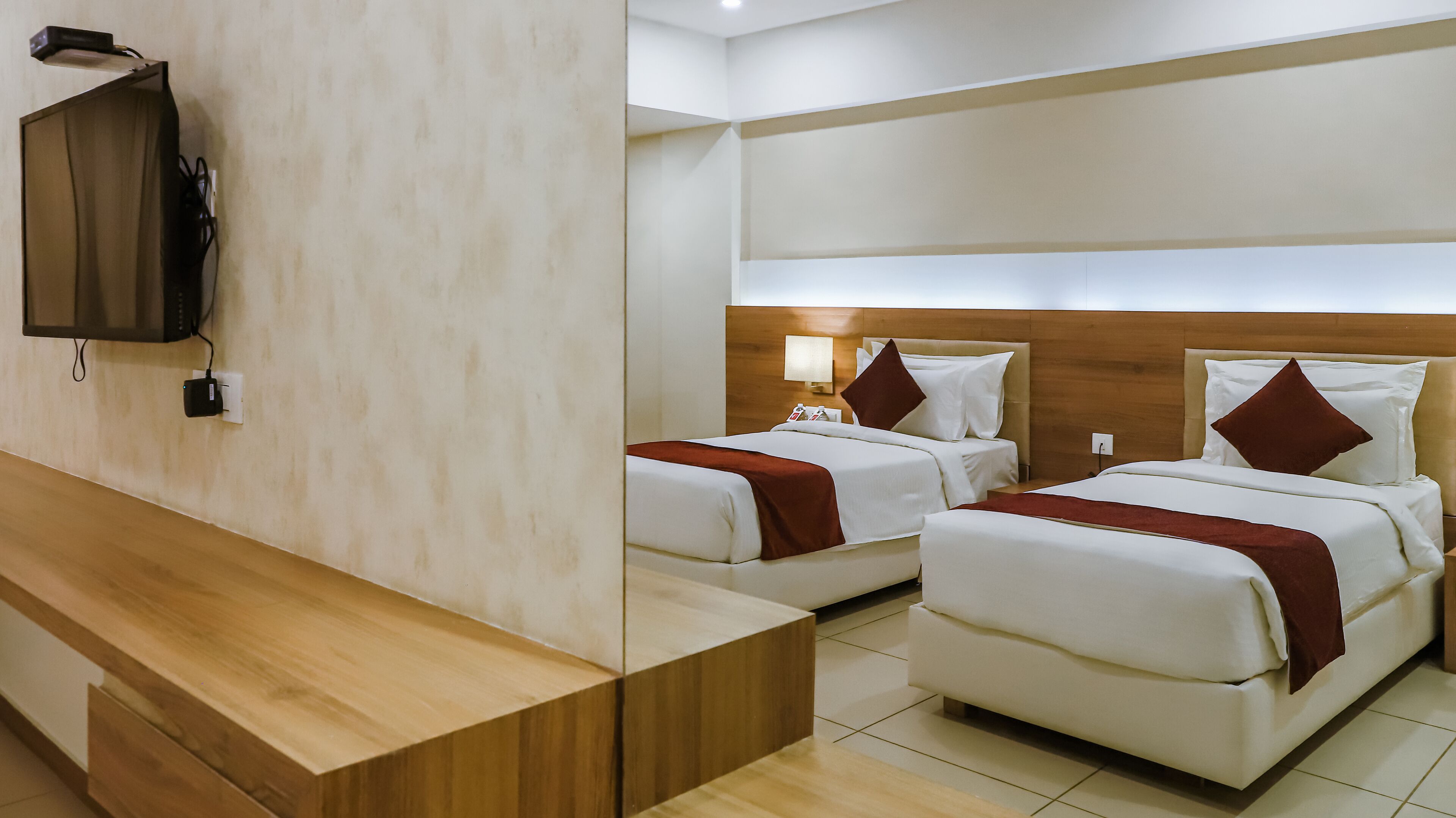 A hotel room with two beds, a wooden bench, a flat-screen TV mounted on the wall, and a bedside table with a lamp.