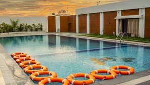 A swimming pool with orange lifebuoys arranged in a line, surrounded by a modern building with a flat roof and a grassy area. - Regenta Central Somnath (Veraval)