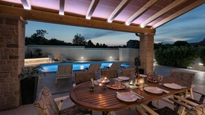Ein Essbereich im Freien mit Pool, Holzmöbeln und einer Pergola mit Beleuchtung.