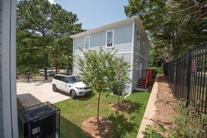 A two-story house with a white SUV parked in the driveway, a small tree in the front yard, and a black metal fence along the side.