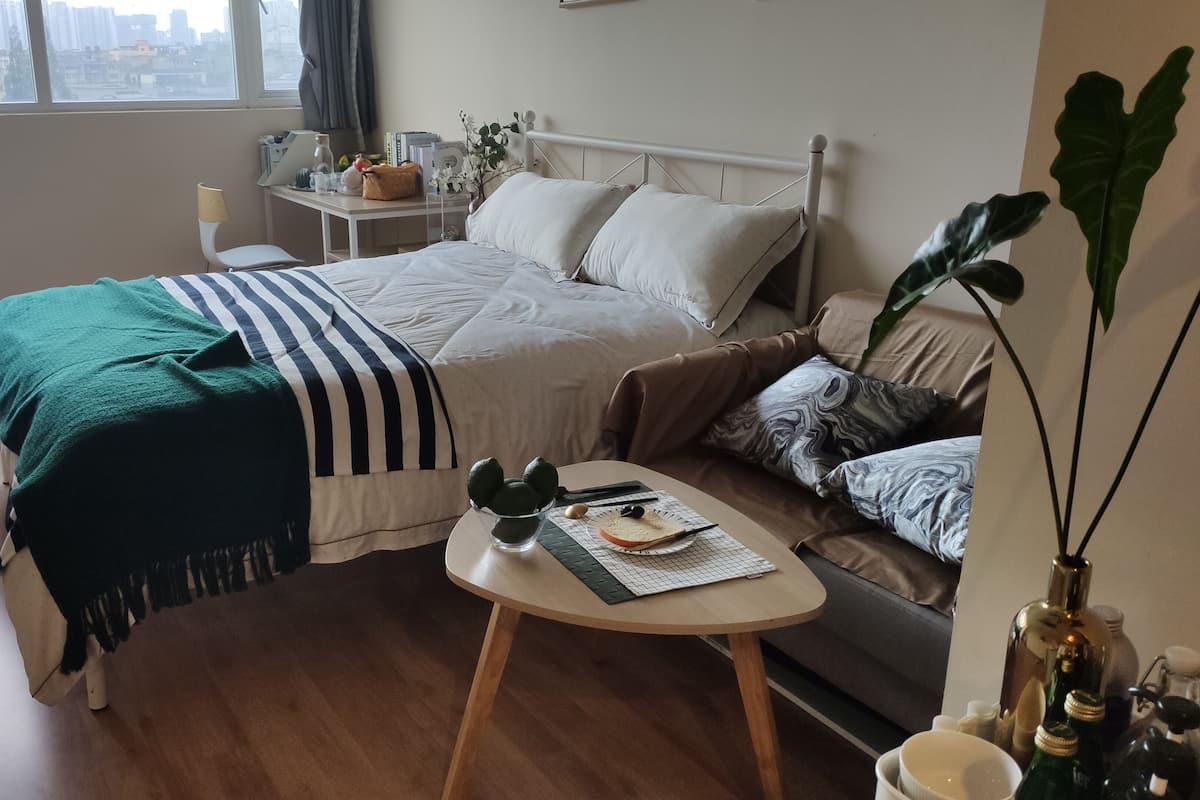 A neatly arranged bedroom with a bed, a sofa, a small table, and a window with a city view.