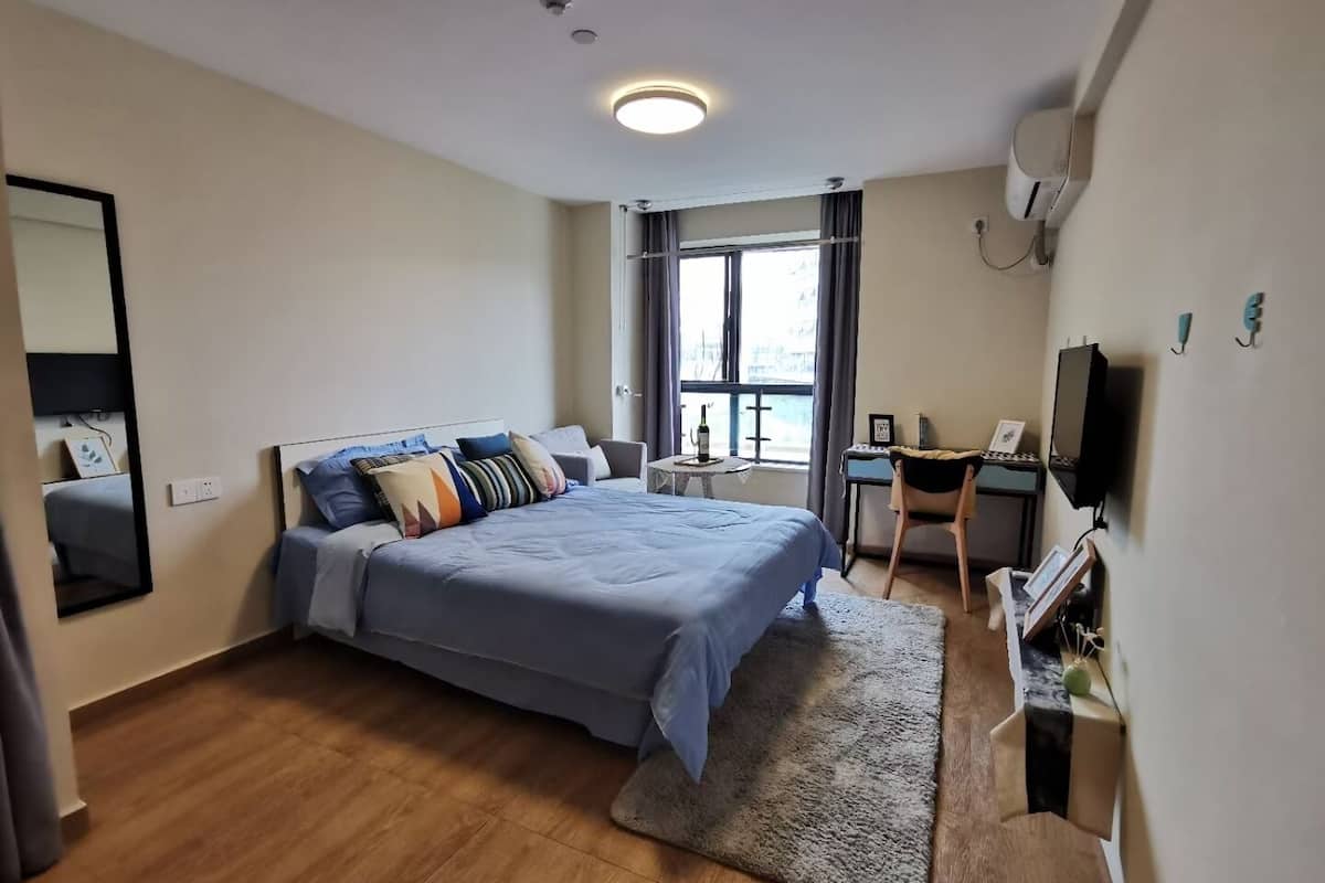 A modern hotel room with a large bed, a desk, a chair, a TV, and a window with a view.