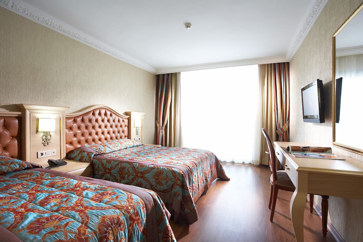 Quarto de hotel com duas camas, uma escrivaninha, uma cadeira, uma televisão e uma janela com cortinas.