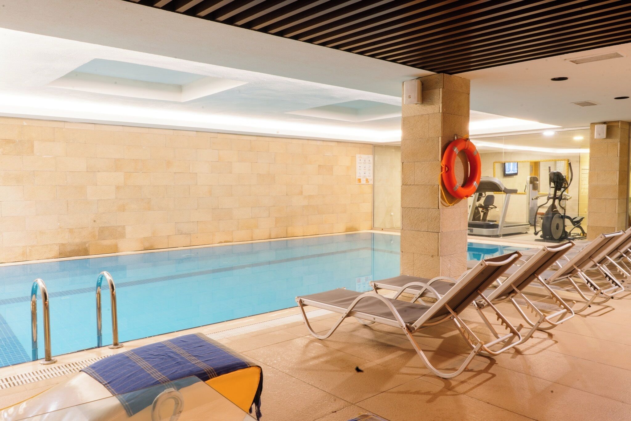 An indoor swimming pool with clear water, lounge chairs, and exercise equipment in the background.