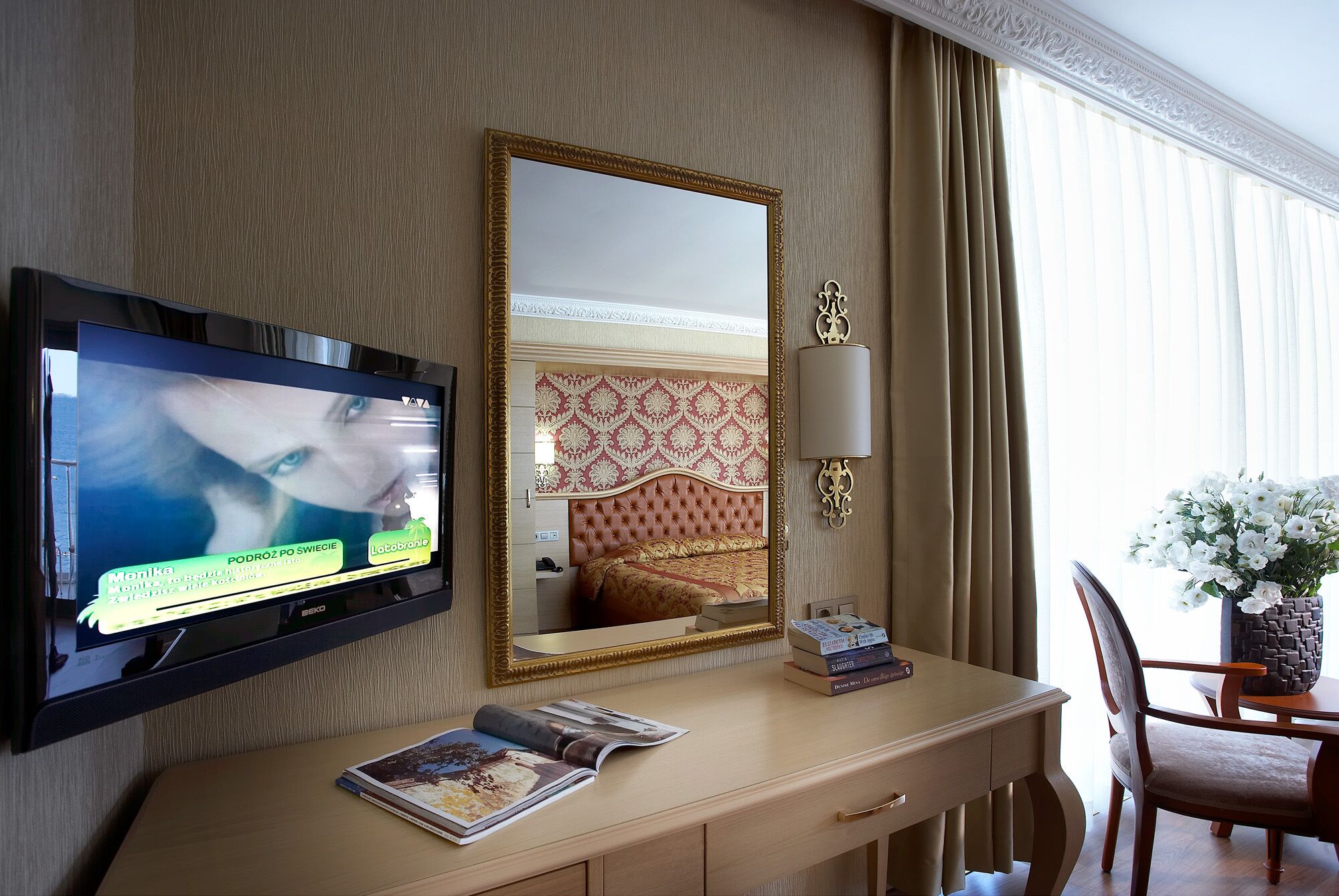 A hotel room with a television, a mirror, a desk with books, a chair, and a vase with flowers.