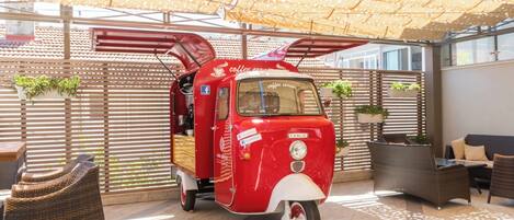 A red vintage van with a coffee theme parked in a covered outdoor area with wicker furniture and potted plants.