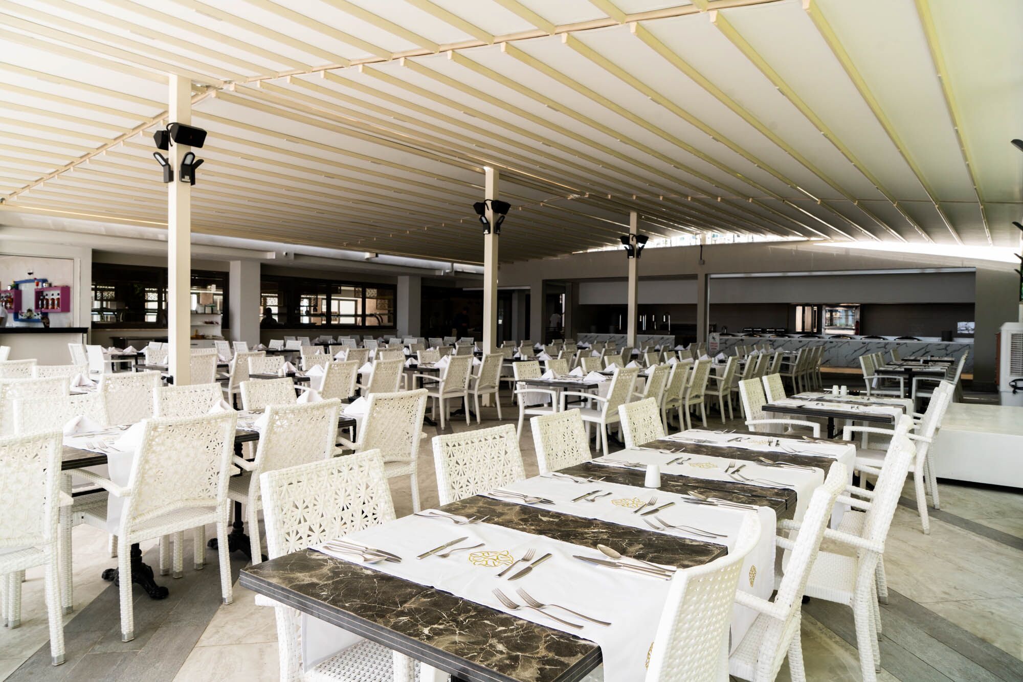 A large indoor dining area with rows of white wicker chairs and tables, a high ceiling with linear lighting, and a view of the kitchen area in the background.