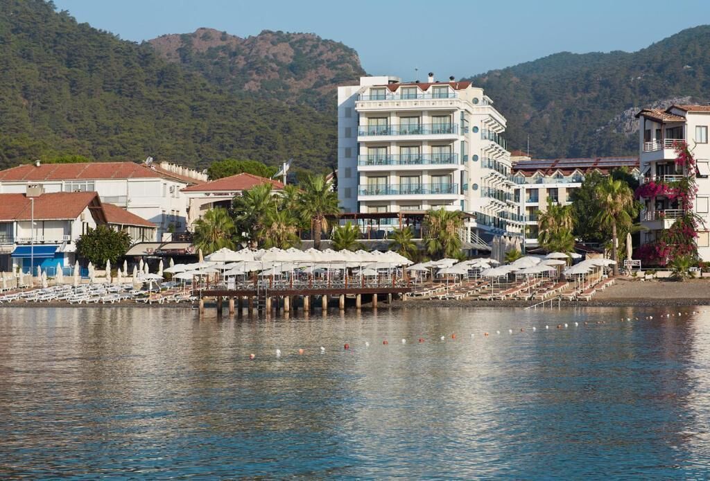 A waterfront hotel with multiple balconies, a pier with umbrellas, and a backdrop of hills.
