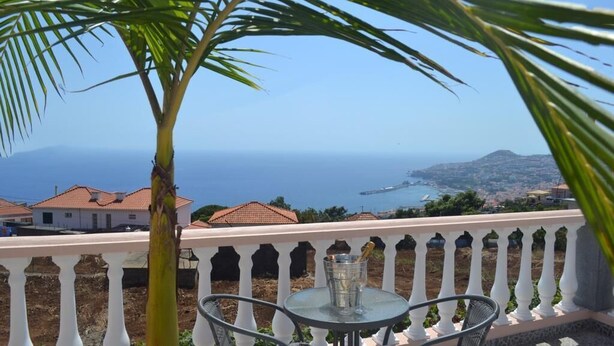 A balcony with a table and chairs, overlooking a coastal view with houses and the sea. - Casa da Paz - Fantastic view of Funchal Bay (São Gonçalo)