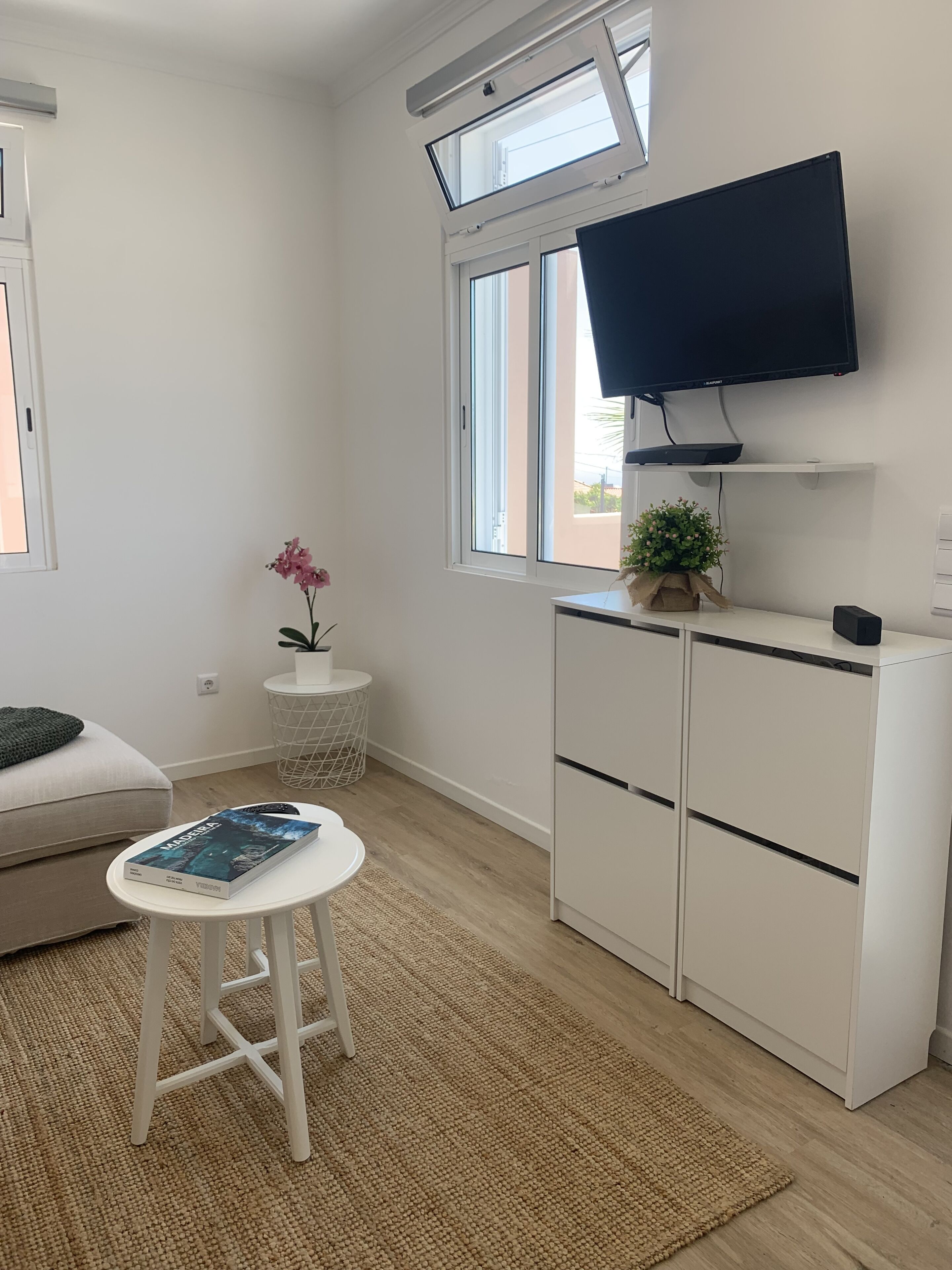 Habitación moderna y luminosa con un ventanal grande, un televisor de pantalla plana en la pared, una mesita redonda con un libro, un sofá blanco y un armario blanco.