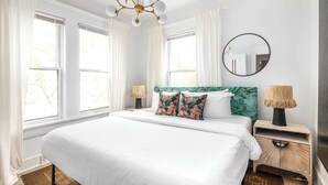 Une chambre d’hôtel avec un grand lit, deux tables de chevet avec des lampes, un miroir rond et une vue sur les arbres par les fenêtres.
