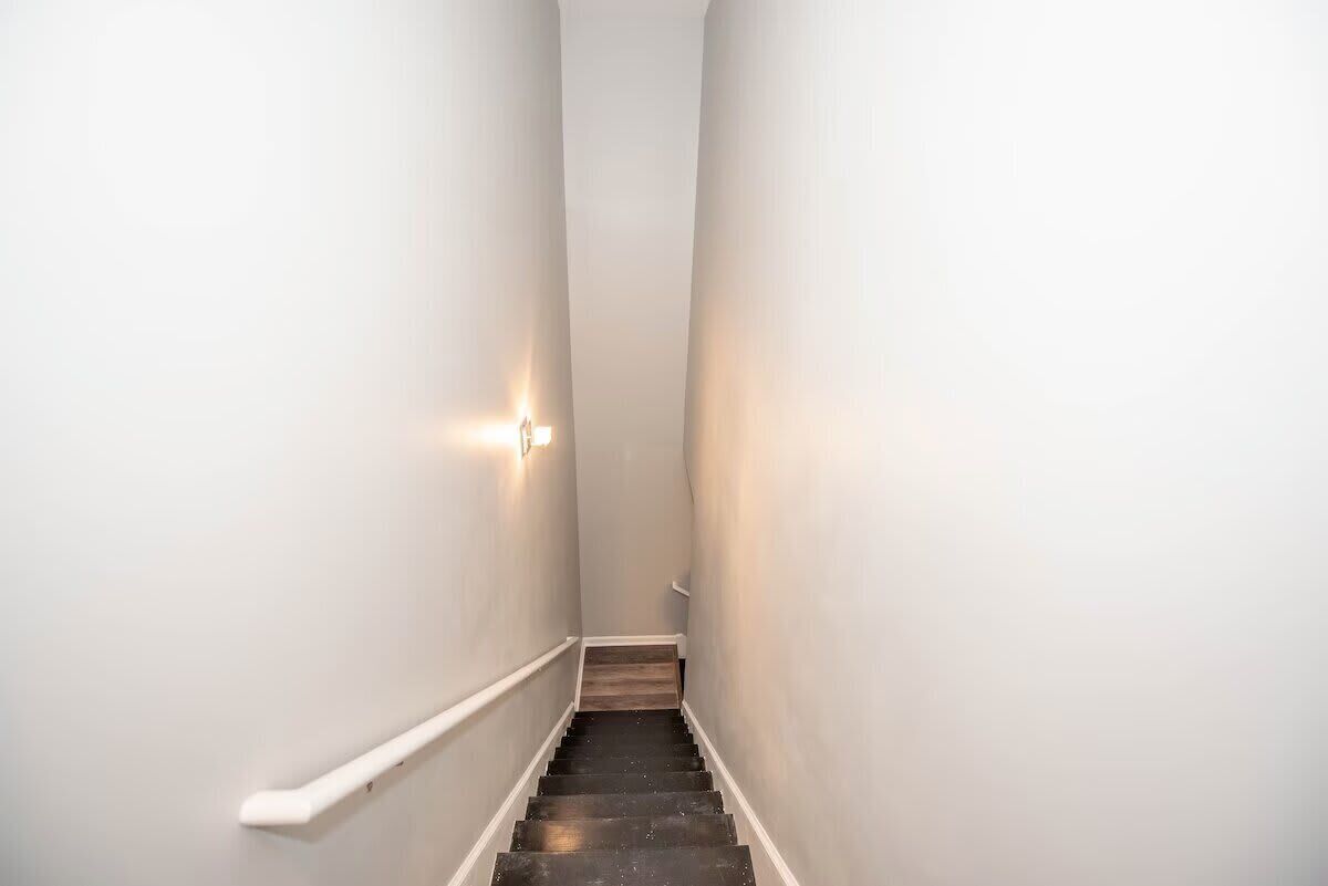 A narrow, white-walled stairway with a wooden handrail and a wall-mounted light fixture.