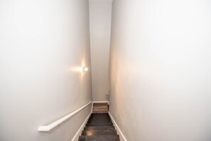 A narrow, white-walled stairway with a wooden handrail and a wall-mounted light fixture.
