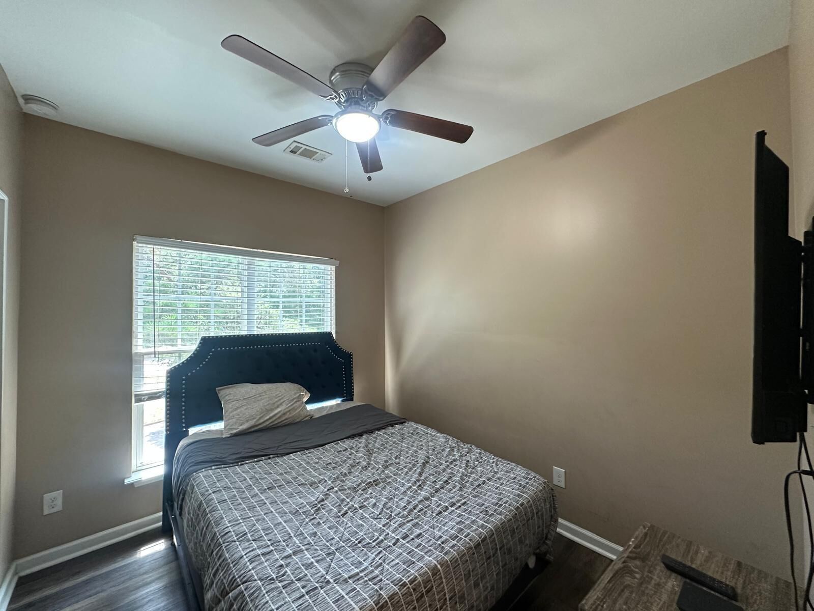 Un dormitorio con cama, ventilador de techo, ventana con persianas y televisor montado en la pared.