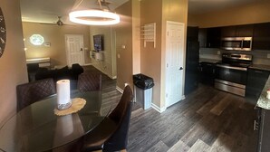 Uma cozinha moderna com eletrodomésticos de aço inoxidável, uma sala de jantar com mesa de vidro e uma sala de estar com televisão.