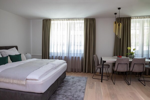 A modern hotel room with a large bed, a dining area with a table and chairs, and large windows with curtains. - Munich Finest Apartments - Studio 5 (München)