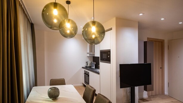 Een moderne eetruimte met een marmeren tafel, drie hanglampen, een televisie en een keuken op de achtergrond.