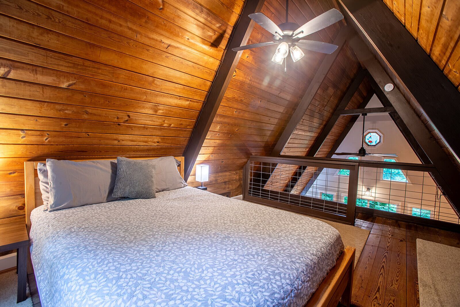A bedroom with a sloped ceiling, a bed with a patterned bedspread, two pillows, a nightstand with a lamp, and a view through a window to the outside.