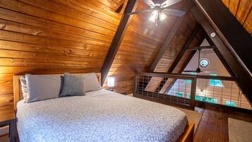A bedroom with a sloped ceiling, a bed with a patterned bedspread, two pillows, a nightstand with a lamp, and a view through a window to the outside.