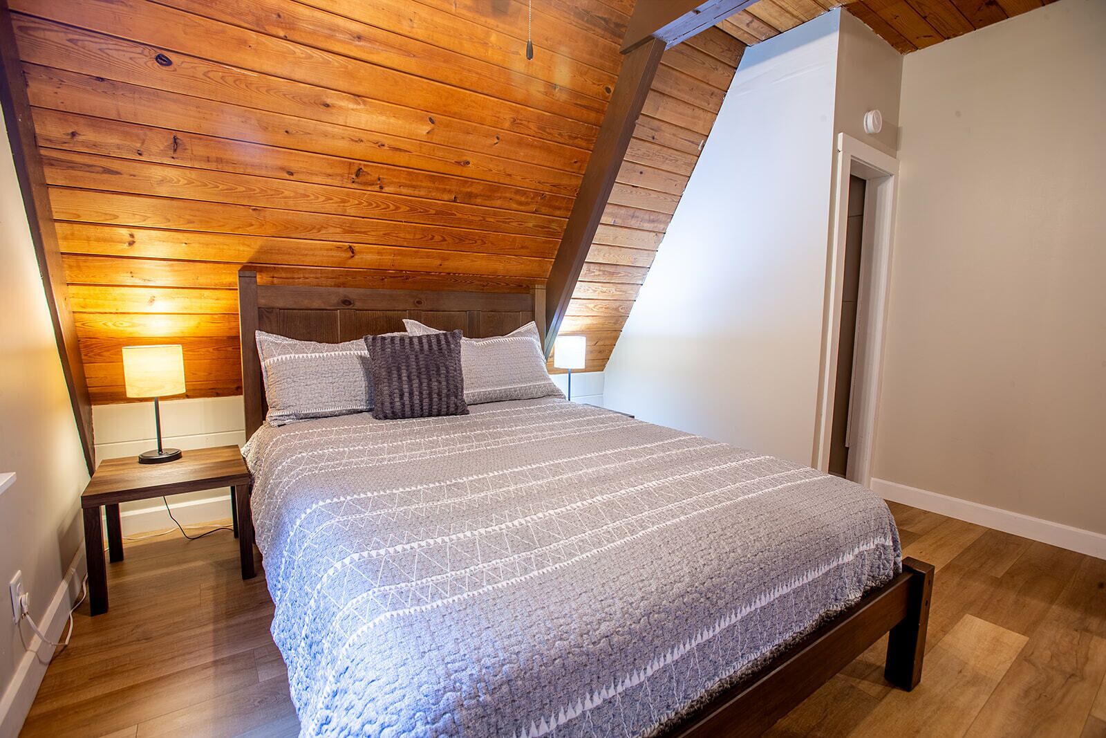 Un dormitorio con techo inclinado, una cama con colcha estampada, dos lámparas de noche y suelo de madera.