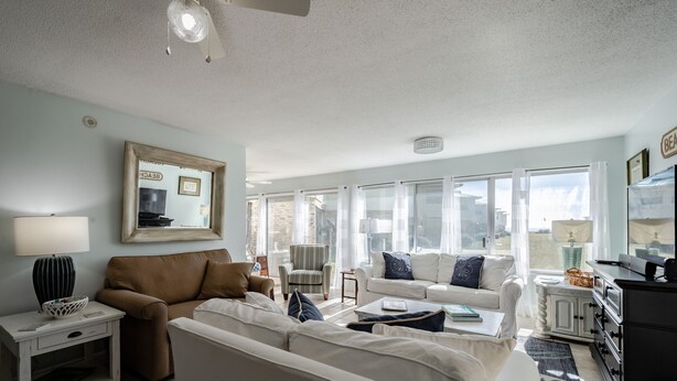 A spacious living room with a brown sofa, white armchairs, and a coffee table. A large mirror hangs on the wall, and there is a window with curtains. - Dauphin Island's East End Jewl (Dauphin Island)
