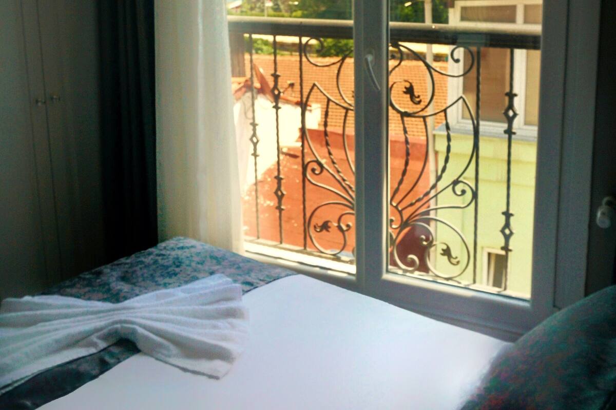 Quarto de hotel com cama, janela com vista para um horizonte urbano e água, e varanda com grade de ferro forjado.