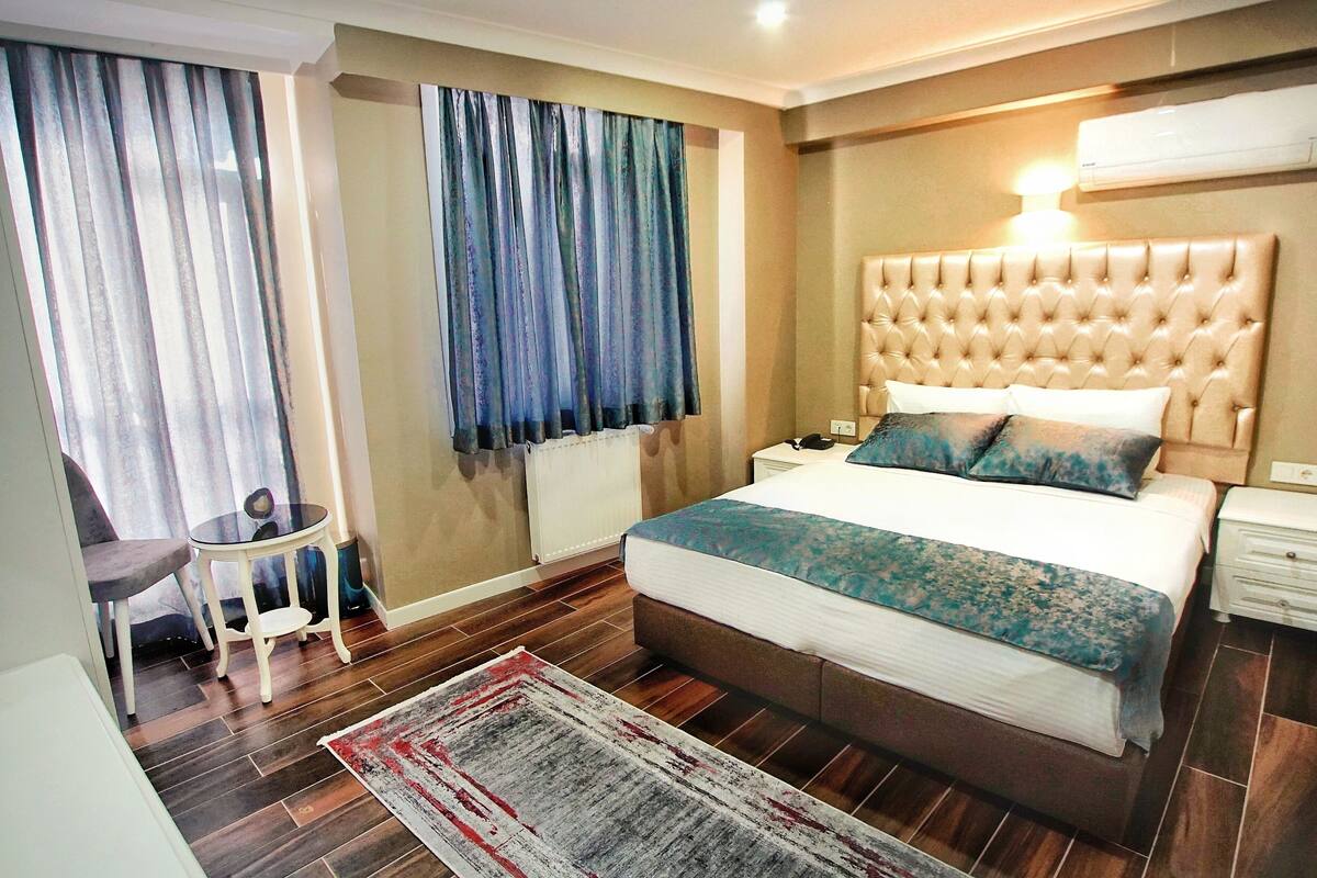 Quarto com cabeceira estofada, cama grande com colcha estampada, mesa de cabeceira com abajur, cadeira e vista para o exterior através de cortinas.
