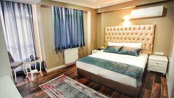 Une chambre à coucher avec une tête de lit capitonnée, un grand lit recouvert d’une couvre-lit à motifs, une table de chevet avec une lampe, un fauteuil et une vue sur l’extérieur à travers les rideaux.